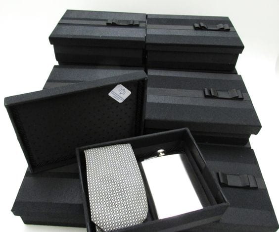 Caixa preta contendo um cantil para bebida em prata e uma gravata cinza com pontos brancos.