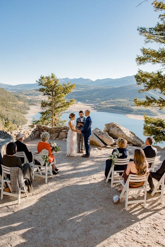 Casamento sendo realizado em uma montanha, com vista para o rio, com alguns poucos convidados sentados em cadeiras brancas.